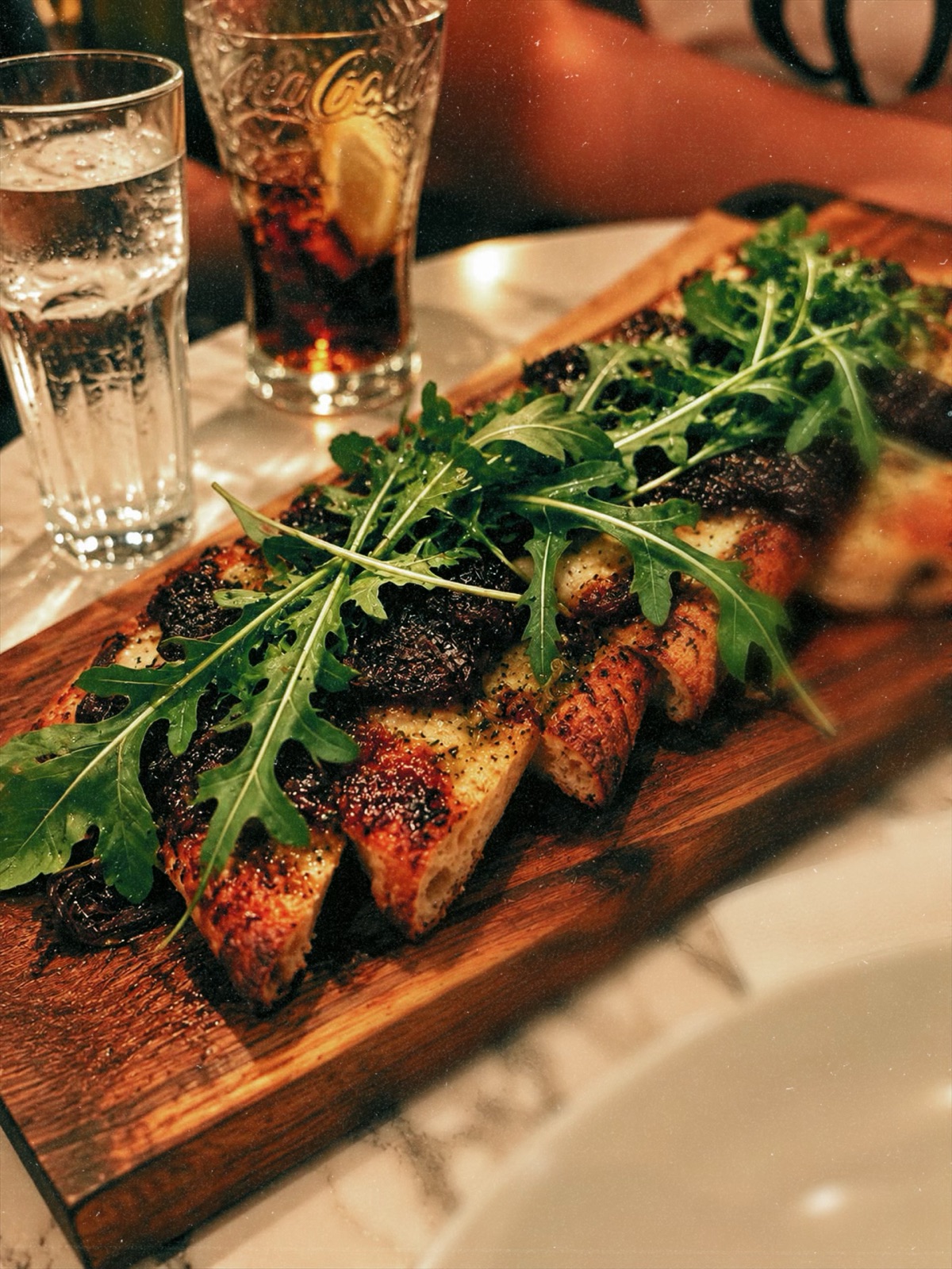 Toasted bread topped with rocket on a wooden board, with drinks alongside