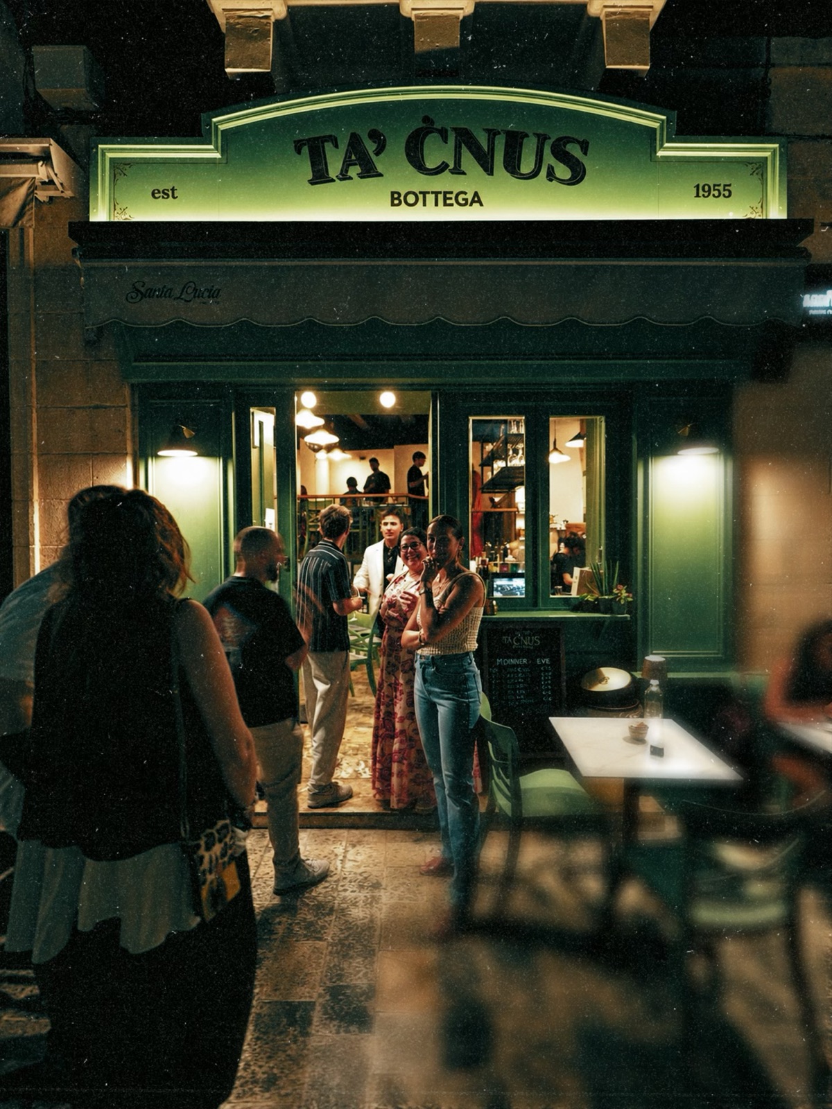 Guests arriving at the lit green shopfront of Bottega Ta’ Ċnus for an evening event